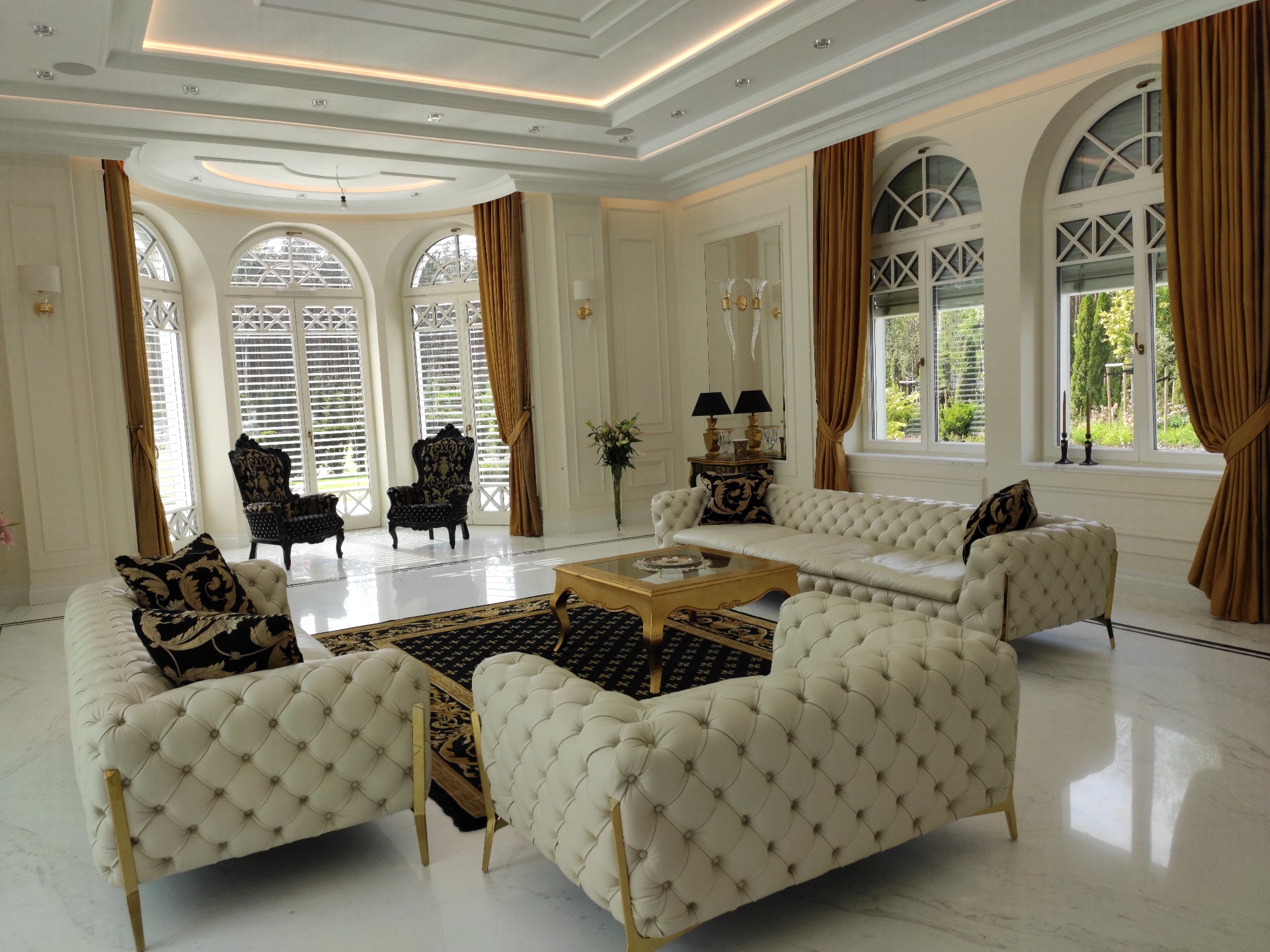 Elegancki salon w stylu glamour z białymi sofami Chesterfield, złotymi akcentami i oknami łukowymi z widokiem na ogród.