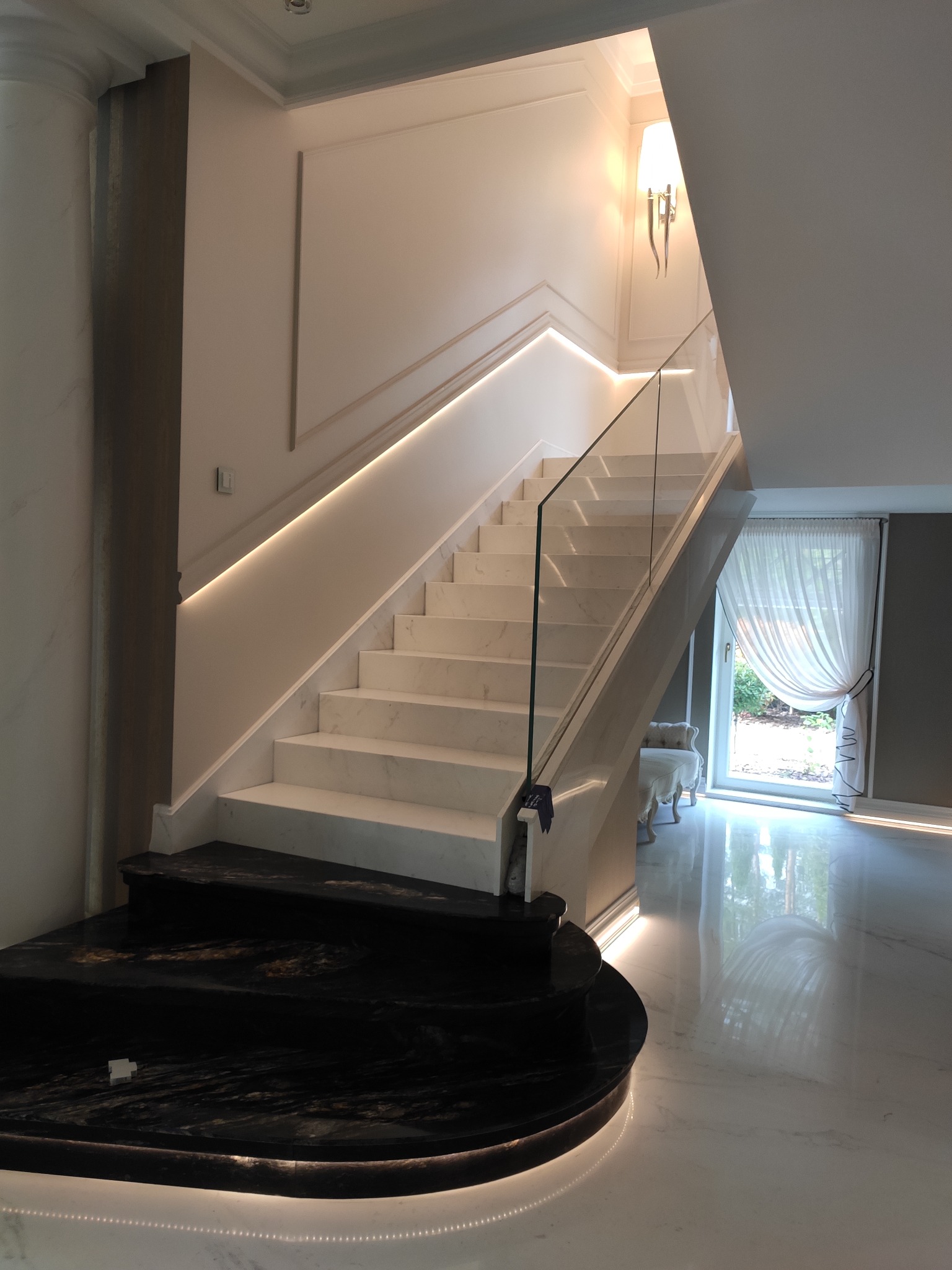 Eleganckie schody z białego marmuru z podświetleniem LED, szklana balustrada, czarny marmurowy stopień początkowy, kinkiet ścienny w stylu retro, widok na okno z firanką.