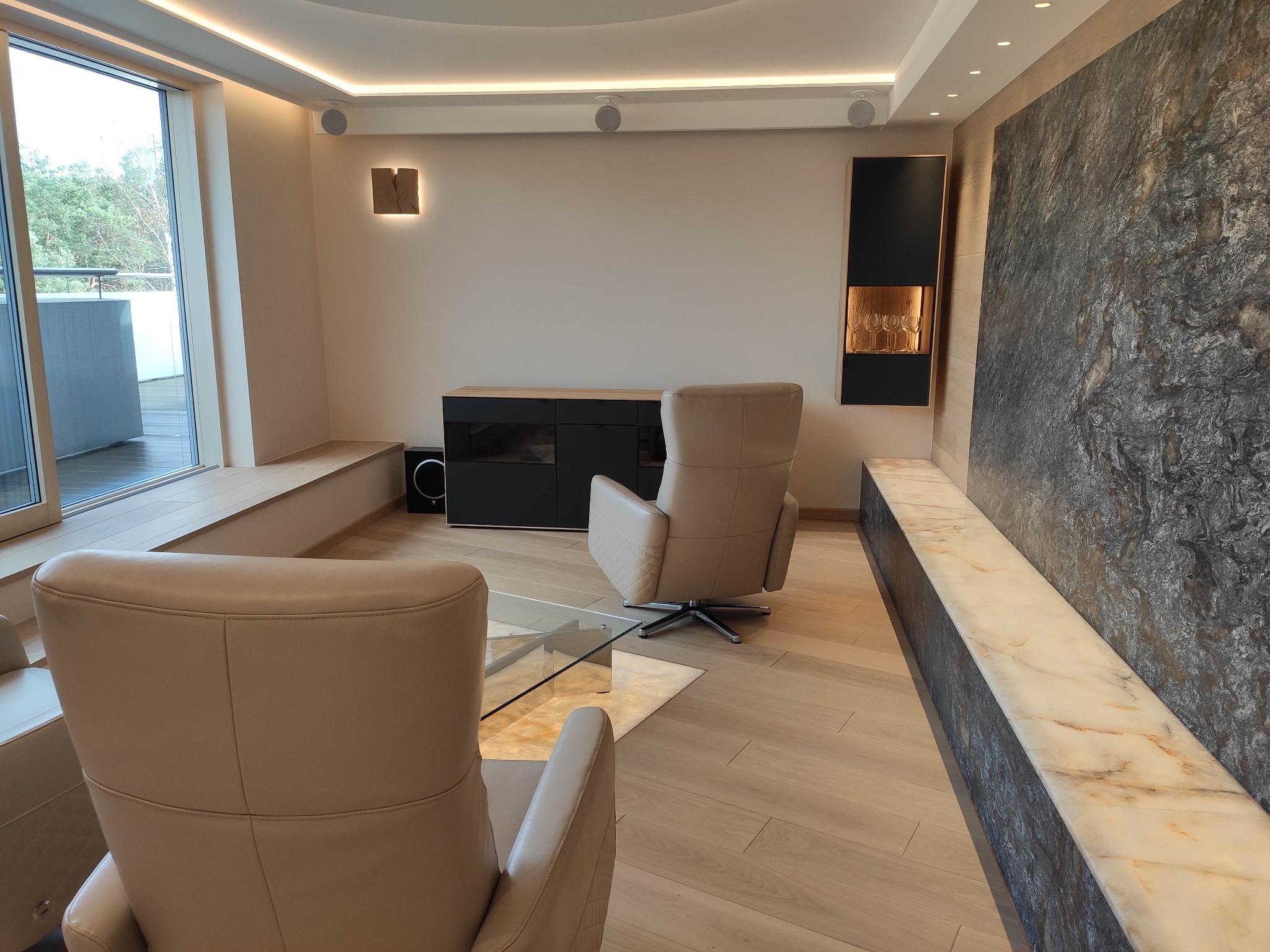 Jasne, nowoczesne wnętrze salonu z dwoma skórzanymi fotelami, minimalistyczną szafką RTV, dekoracyjną ścianą z kamienia i oświetleniem punktowym LED w suficie.