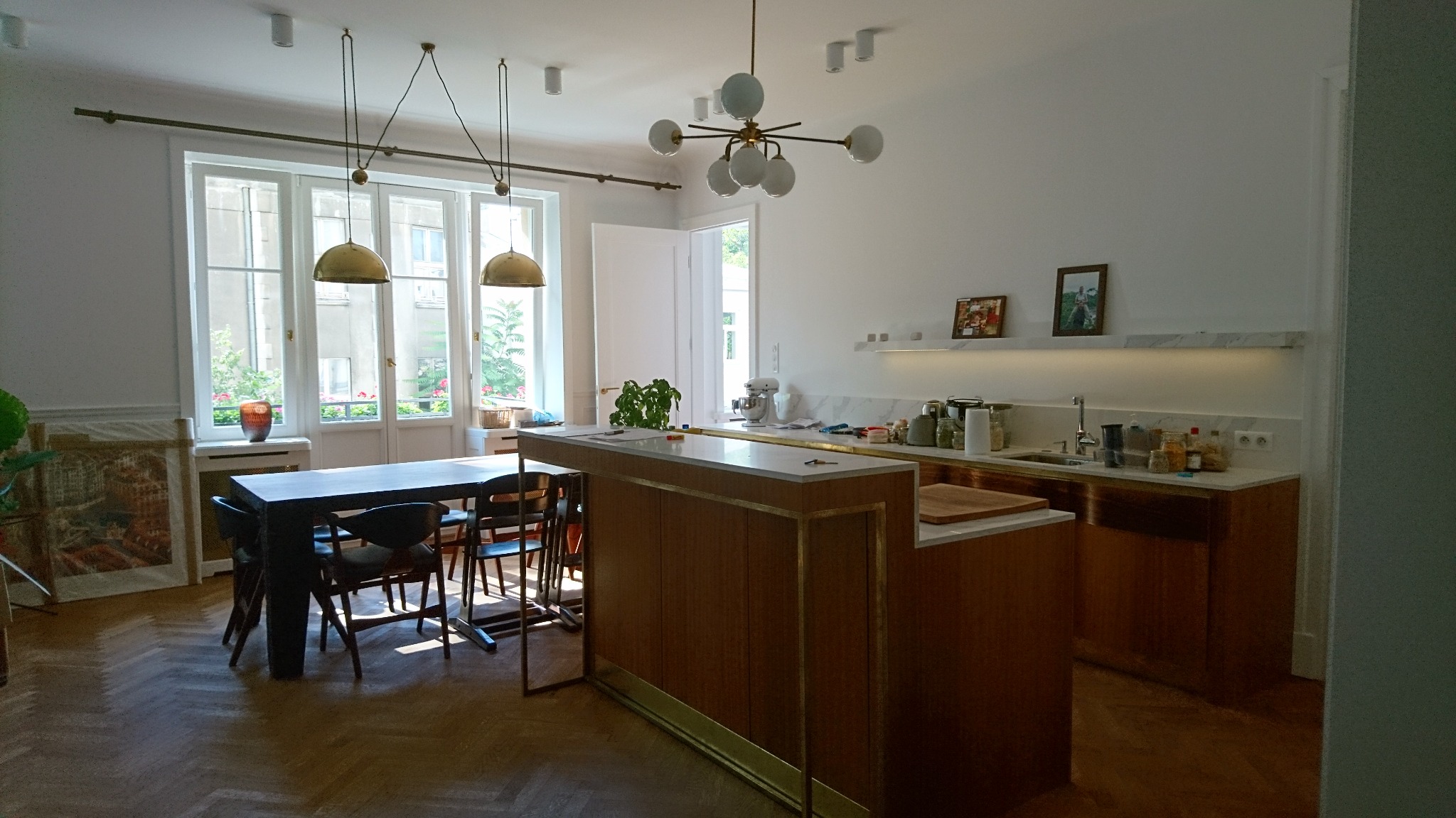 Jasne, przestronne wnętrze kuchni z wyspą, oświetlone wiszącymi lampami w stylu retro i nowoczesnym, z widokiem na zieleń za oknem.