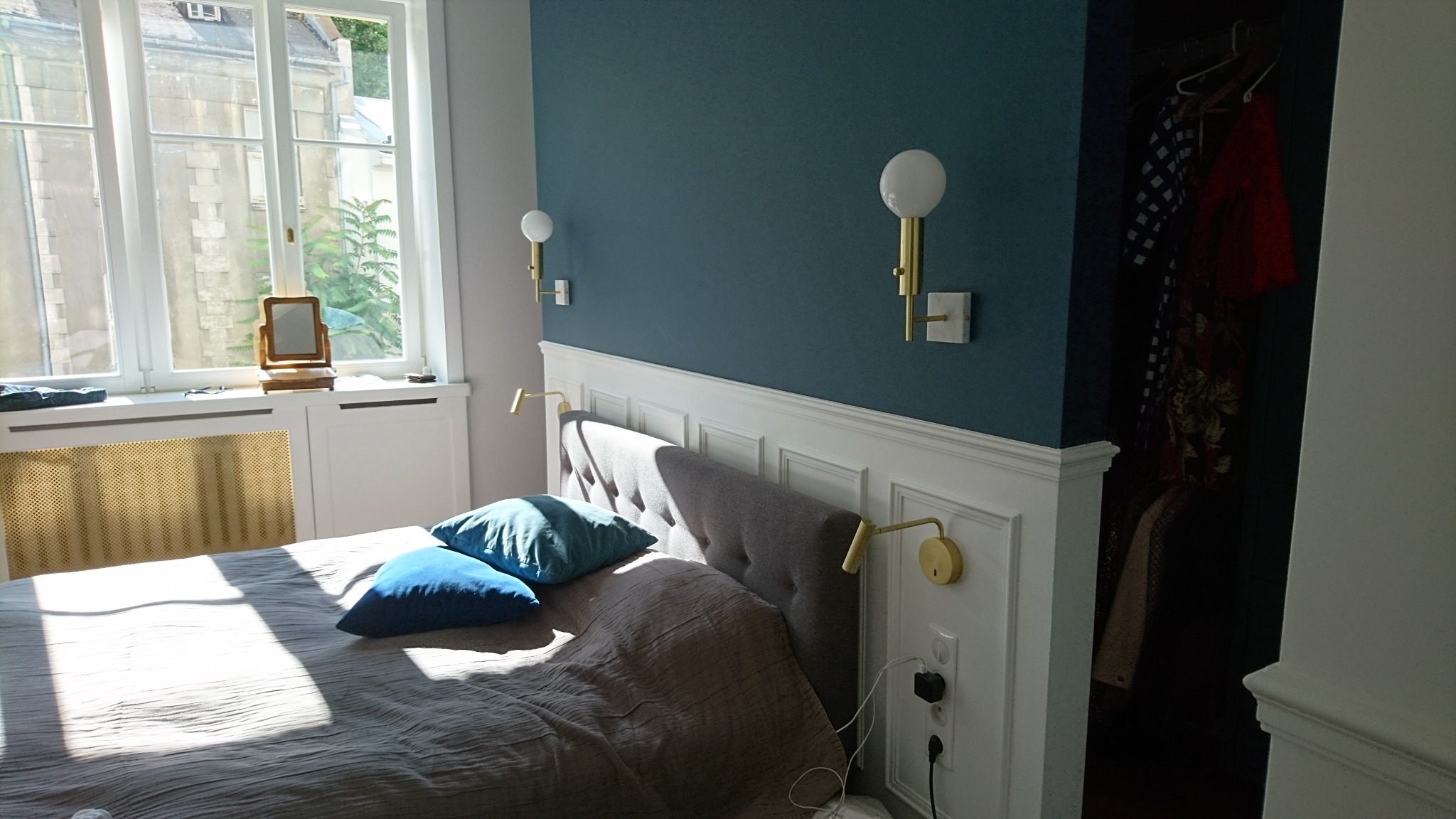 Sypialnia z łóżkiem, dwiema poduszkami, lampkami ściennymi z białymi kloszami i gniazdkiem elektrycznym z podłączonym zasilaczem, oświetlona promieniami słońca.