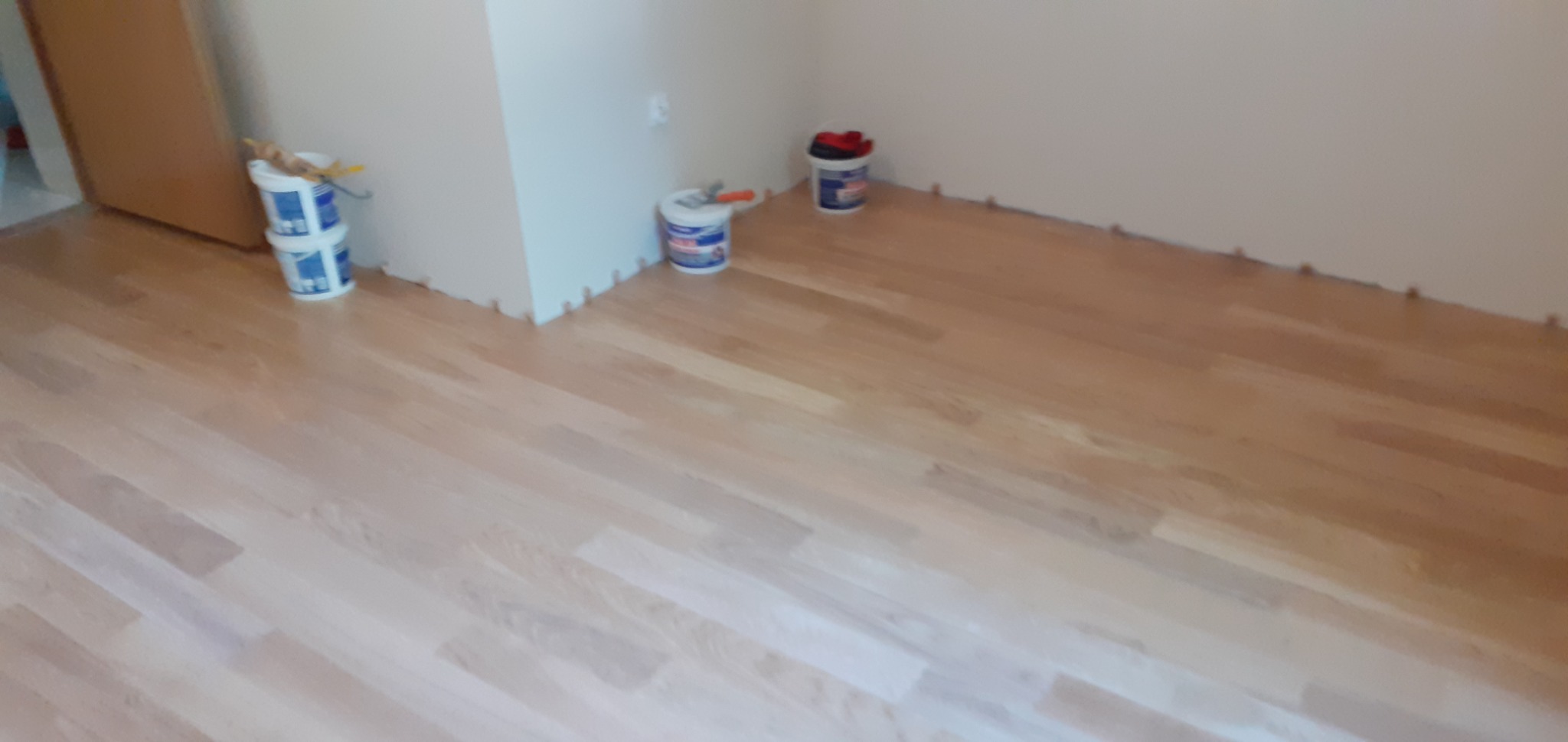 Świeżo polakierowana, jasna podłoga drewniana w pokoju, z widocznymi puszkami z farbą i narzędziami malarskimi przy ścianach, zabezpieczona listwami przypodłogowymi w trakcie prac wykończeniowych.