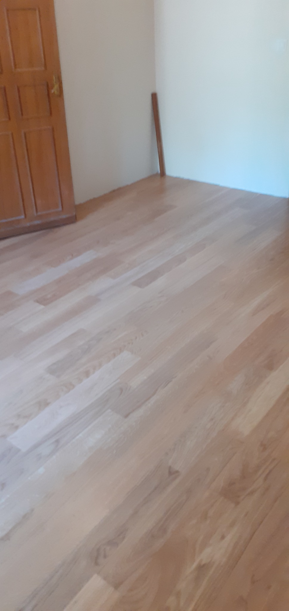 Jasna, świeżo ułożona podłoga z drewnianych paneli, widoczna przy ścianie i brązowych drzwiach w jasnym wnętrzu.