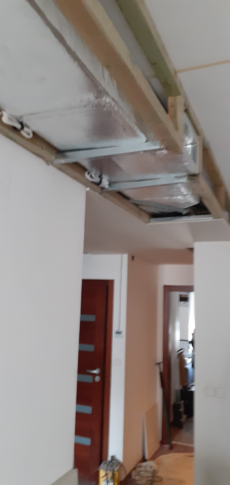 Montaż sufitu napinanego: widoczne drewniane ramy i folia izolacyjna w trakcie instalacji, z perspektywy pomieszczenia z dwiema parami drzwi w tle.