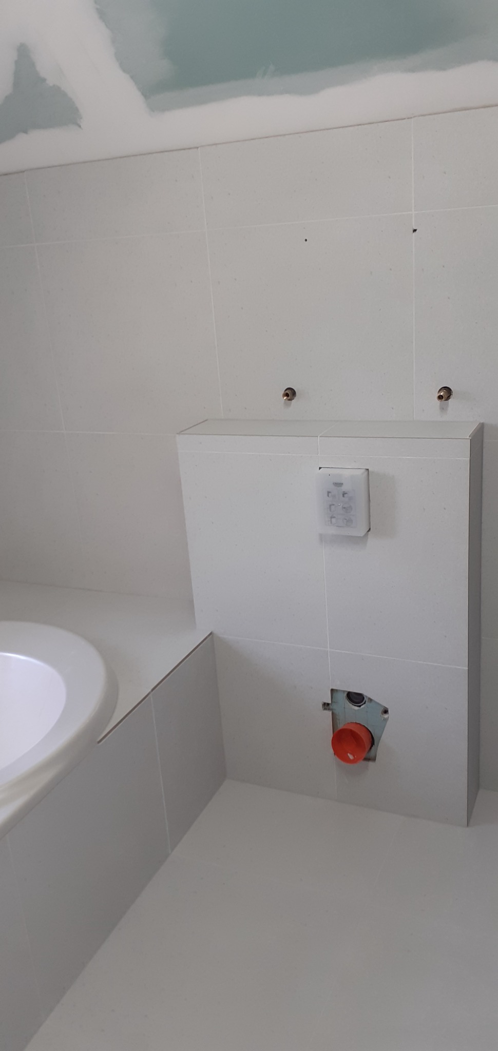 Biała łazienka w trakcie wykańczania: obudowa stelaża WC z zamontowanym przyciskiem spłukującym, widoczne fragmenty wanny i sufitu z płyt karton-gips, niezamaskowane przyłącza wodne.