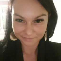 Portret uśmiechniętej kobiety z ciemnymi włosami i dużymi, złotymi kolczykami, ubrana w czarną marynarkę.