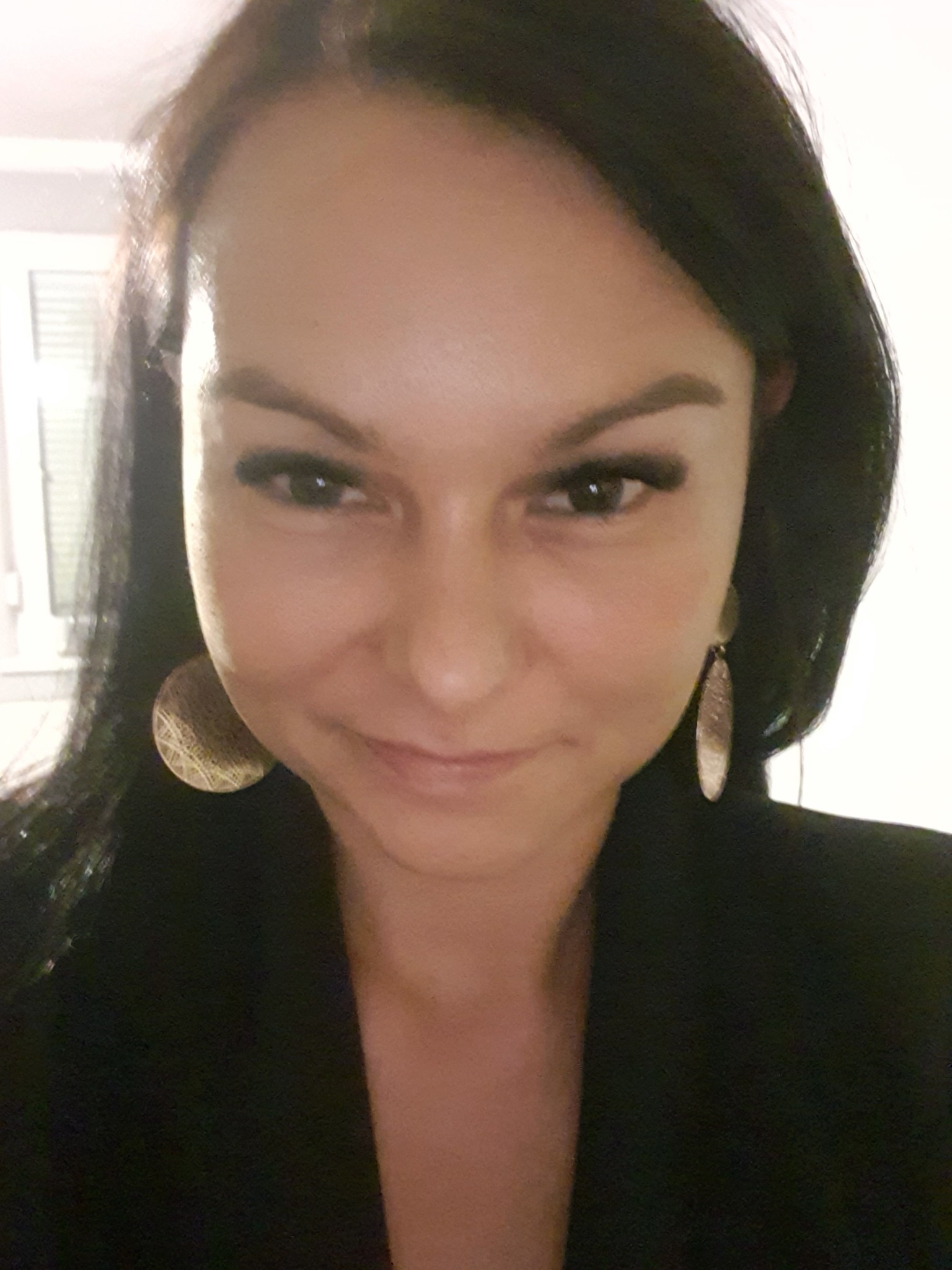 Portret uśmiechniętej kobiety z ciemnymi włosami i dużymi, złotymi kolczykami, ubrana w czarną marynarkę.