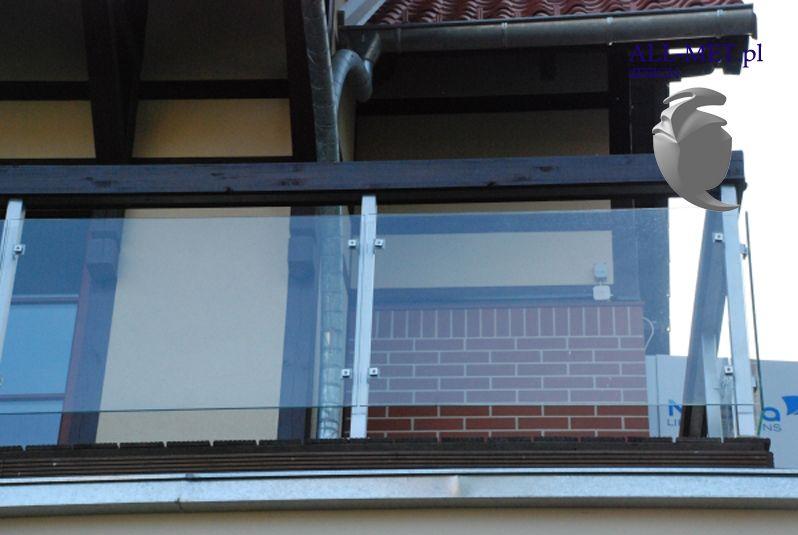Szklana balustrada balkonowa z elementami ze stali nierdzewnej, widok na ceglany fragment ściany i okap dachu.