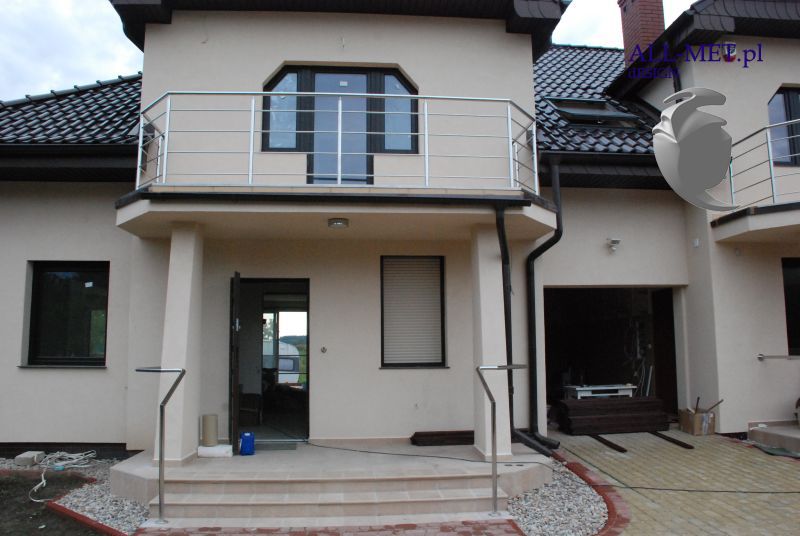 Dwukondygnacyjny dom z beżową elewacją, balkonem z balustradą ze stali nierdzewnej i poręczą przy schodach wejściowych, wykonane przez All-met Olsztyn.