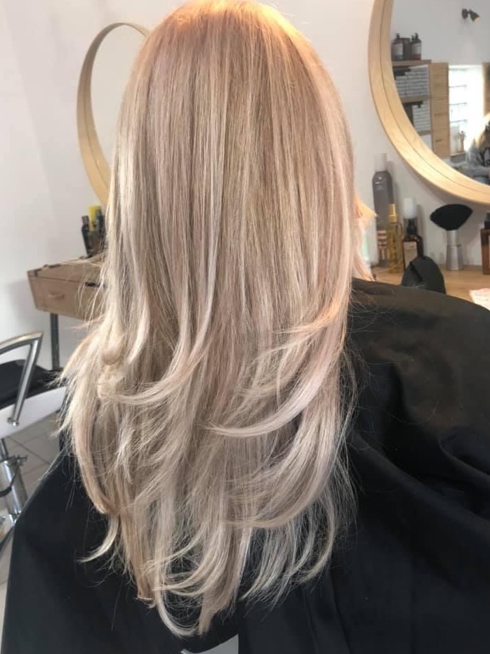 Długie, warstwowe włosy w odcieniach blondu, widok z tyłu, z delikatnym cieniowaniem i refleksami, w tle okrągłe lustro i kosmetyki na półce.