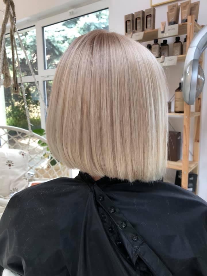 Tył głowy kobiety z krótkim, prostym blond bobem; widoczny czarny fartuch ochronny i półka z kosmetykami w tle.