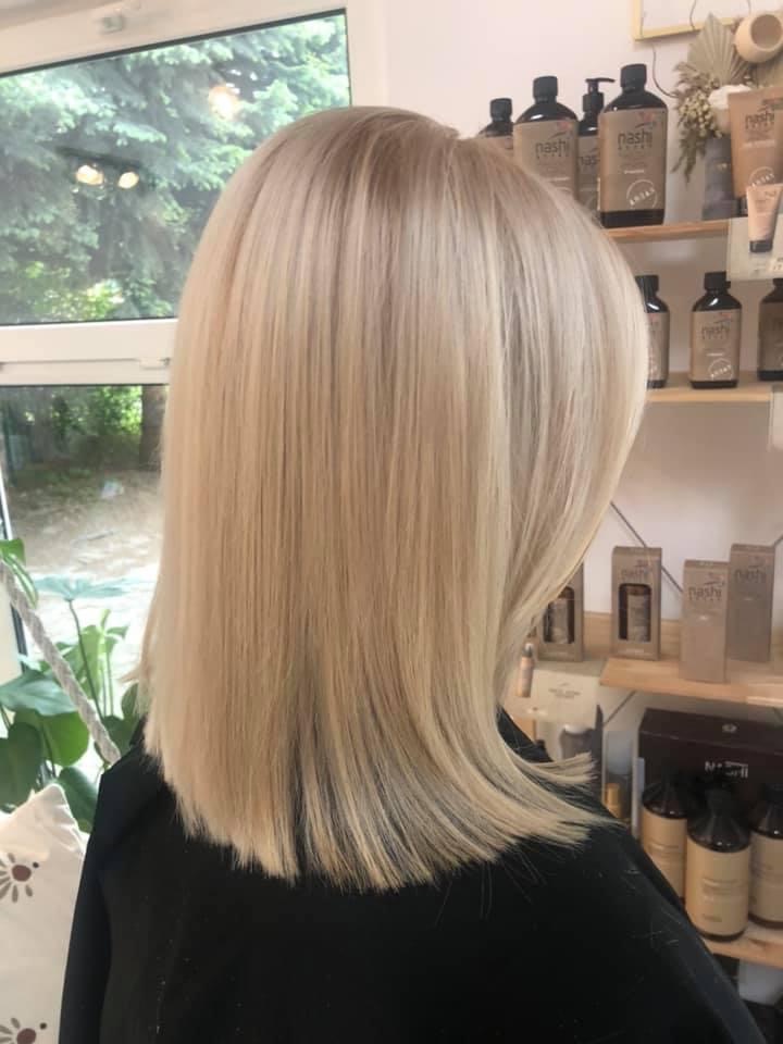 Blond włosy do ramion, świeżo po koloryzacji i strzyżeniu, z widocznymi kosmetykami do pielęgnacji włosów marki Nashi na półce w tle.