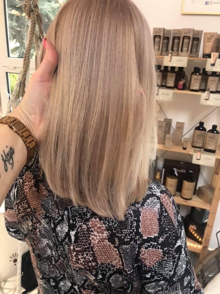 Klientka z prostymi, blond włosami po koloryzacji w salonie fryzjerskim, widoczna od tyłu, z półkami produktów do pielęgnacji włosów w tle.