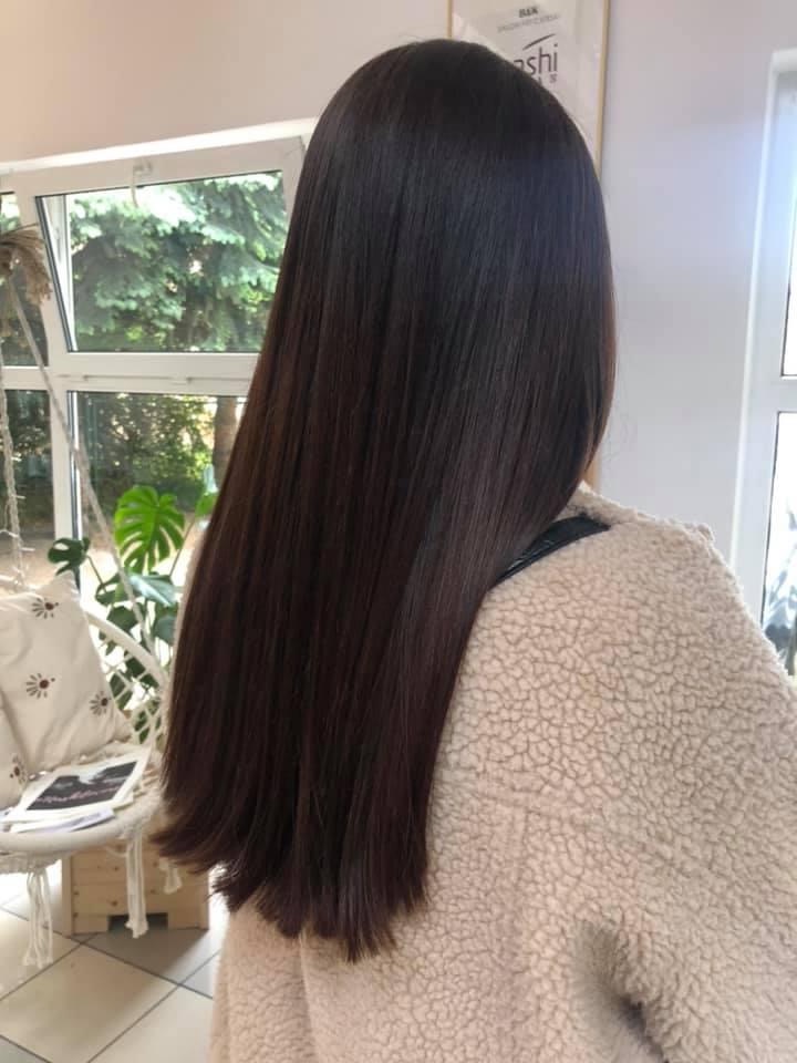 Długie, proste, ciemne włosy z delikatnym połyskiem, widoczne od tyłu, na tle jasnego wnętrza salonu z roślinami i białymi meblami.