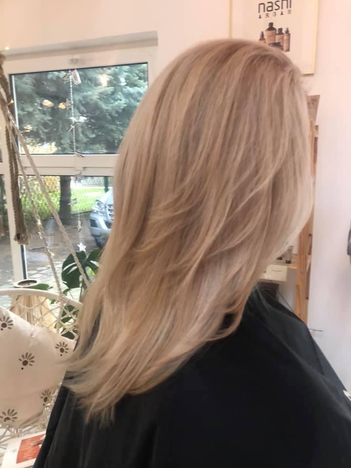 Świeżo ostrzyżone, długie, warstwowe blond włosy, widoczne z profilu, z delikatnymi refleksami, na tle okna i plakatu z produktami do pielęgnacji włosów.