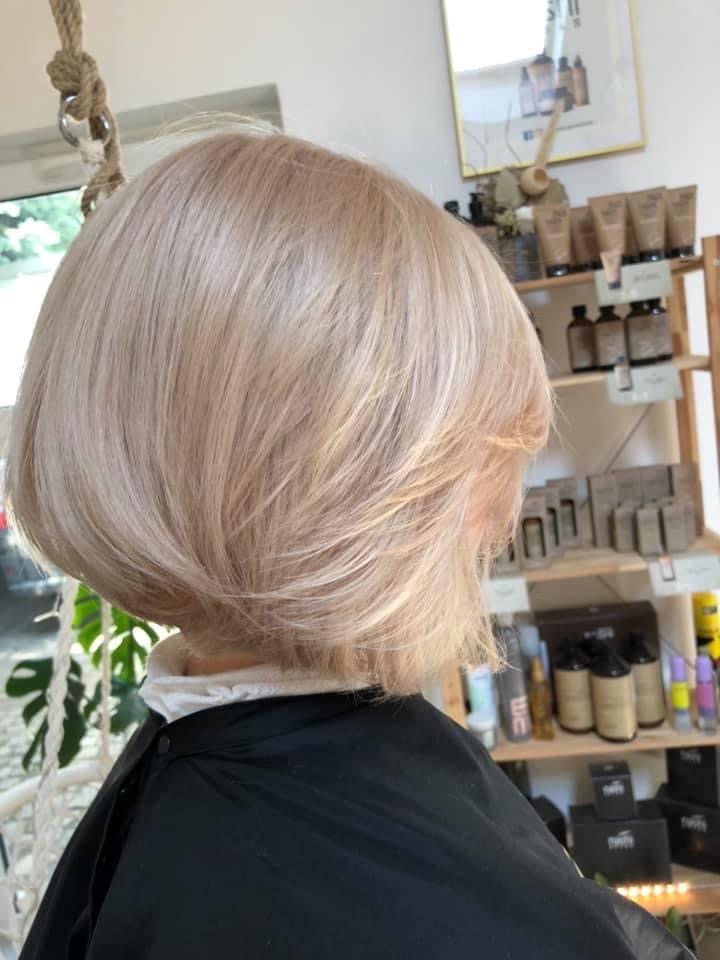 Jasny blond bob z delikatnymi warstwami, widok z tyłu, czarna peleryna ochronna, półki z kosmetykami w tle.