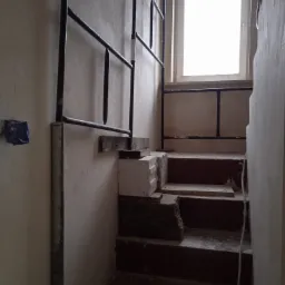 Schody w trakcie remontu z widocznym rusztowaniem, surowymi ścianami i niebieską puszką elektryczną.