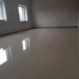 Świeżo wylana, gładka i błyszcząca wylewka anhydrytowa w jasnym pomieszczeniu z dwoma oknami, odbijającymi światło w powierzchni posadzki.