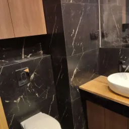 Nowoczesna łazienka z czarnymi marmurowymi płytkami, podwieszaną toaletą z chromowanym przyciskiem spłukiwania, drewnianą szafką pod umywalką nablatową i minimalistycznym oświetleniem.