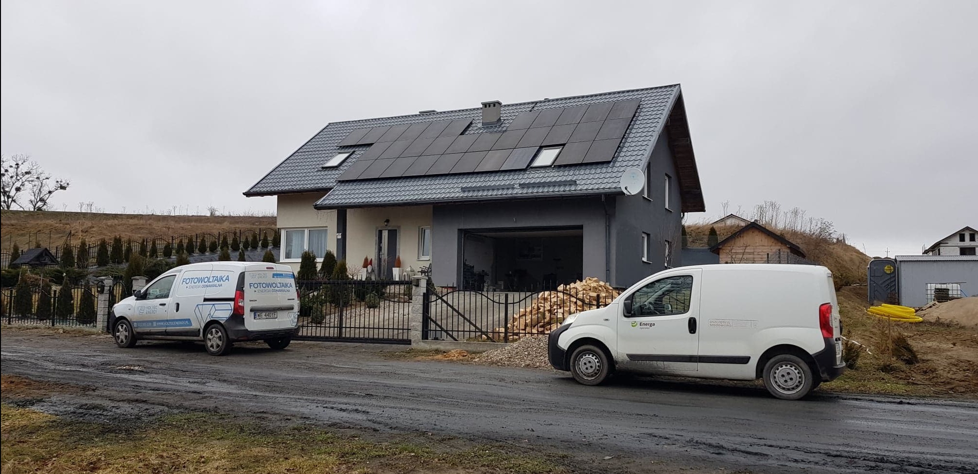 Dom jednorodzinny z zamontowanymi panelami słonecznymi na dachu, na podjeździe zaparkowane dwa samochody dostawcze z logotypami firm. Jeden z nich ma wyraźny napis 'Fotowoltaika energia odnawialna'.
