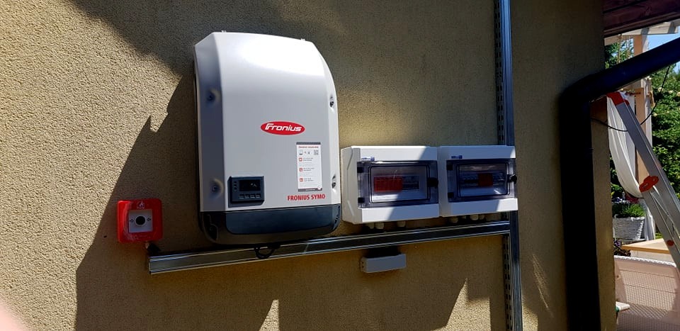 Zamontowany inwerter Fronius Symo wraz z dwoma rozdzielnicami na elewacji budynku, widoczny również czujnik pożarowy i fragment drabiny.