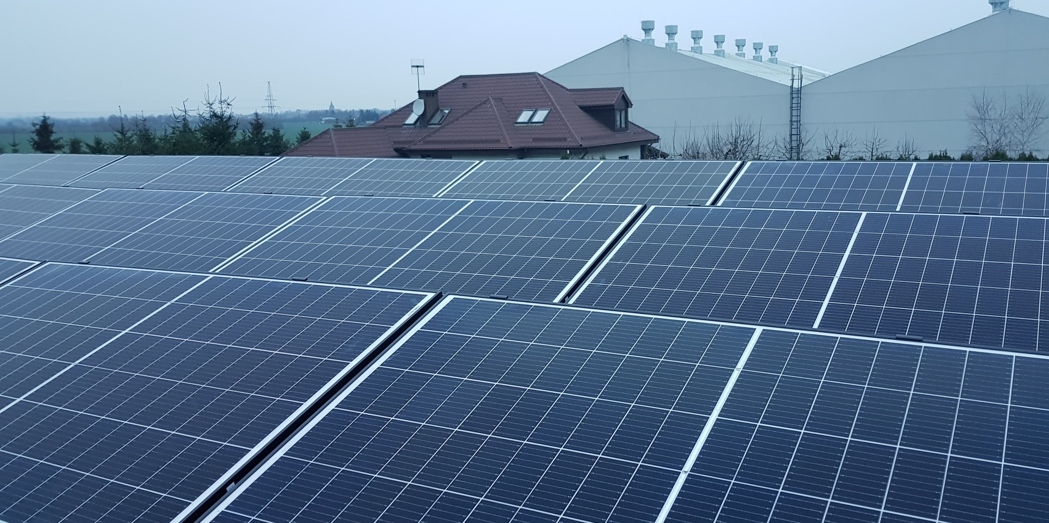 Instalacja paneli słonecznych na dachu, w tle dom jednorodzinny i budynek przemysłowy, pochmurne niebo.