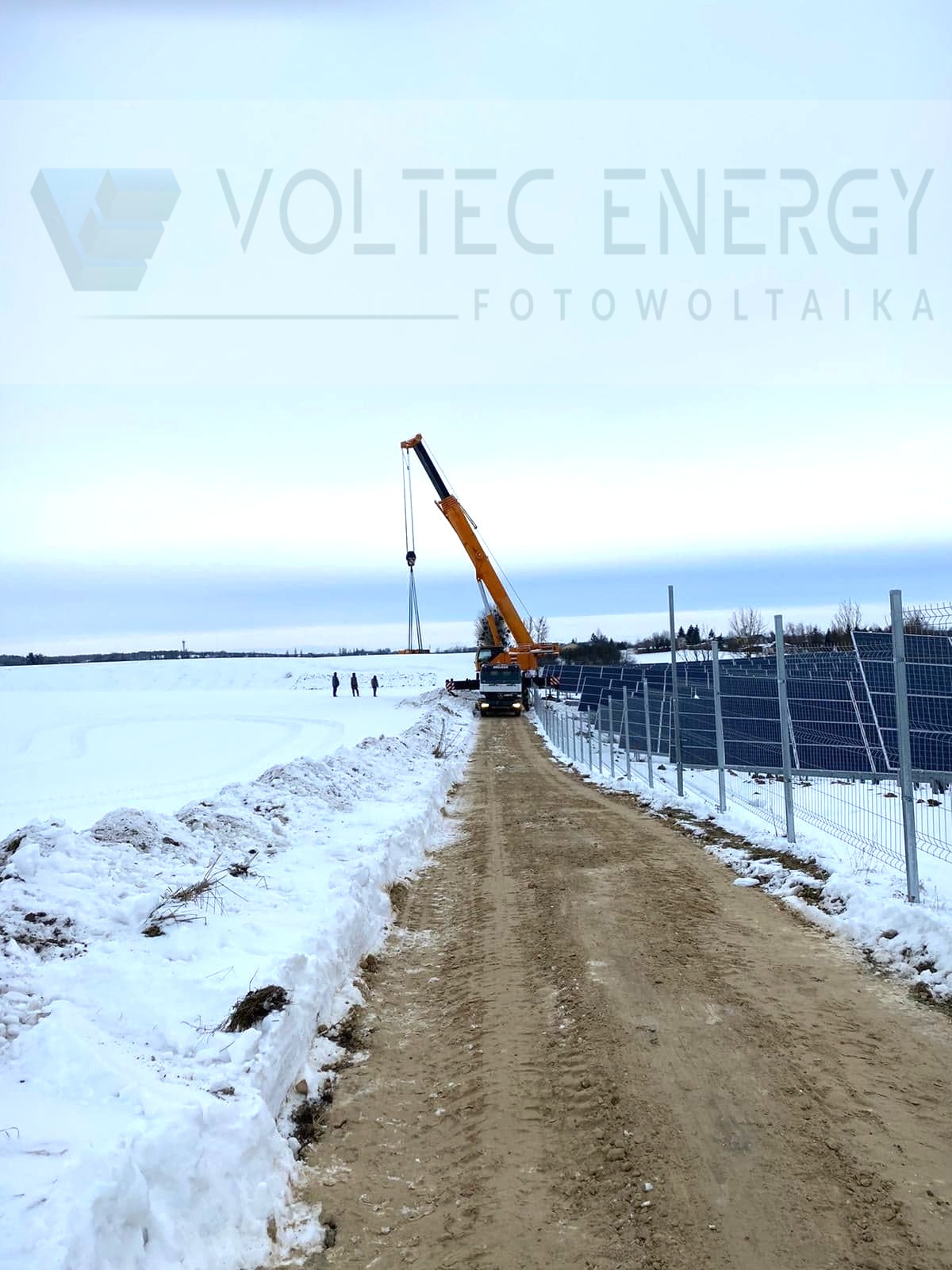 Montaż paneli słonecznych zimą: dźwig podnosi panel na tle zasnieżonego krajobrazu, widoczni pracownicy i częściowo zainstalowane panele, logo firmy Voltec Energy w górnej części kadru.