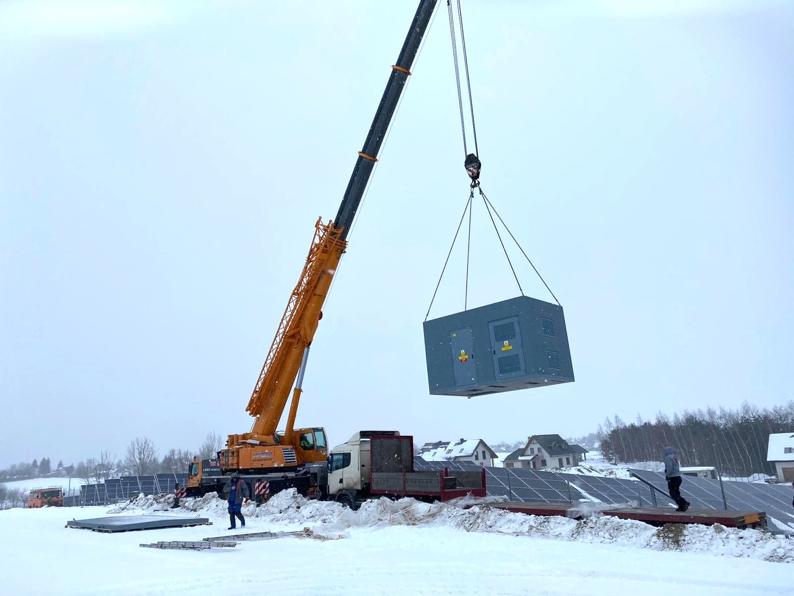 Żuraw przenosi dużą szarą skrzynię z panelami elektrycznymi na tle instalacji paneli słonecznych pokrytych śniegiem, widoczne domy w oddali.