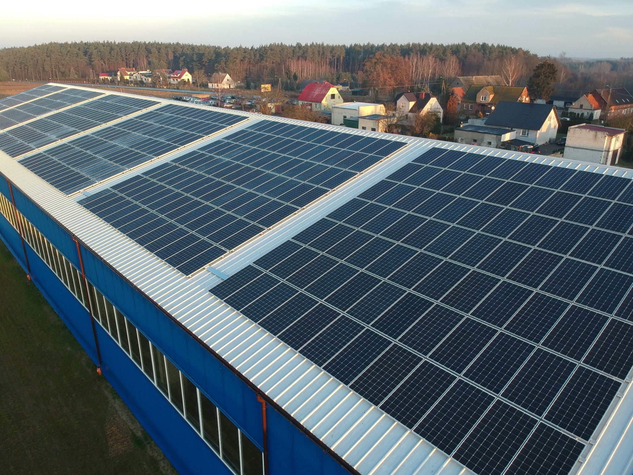Rozległa instalacja paneli słonecznych na dachu niebieskiego budynku przemysłowego, widoczna z góry, w tle zabudowania wiejskie i las.