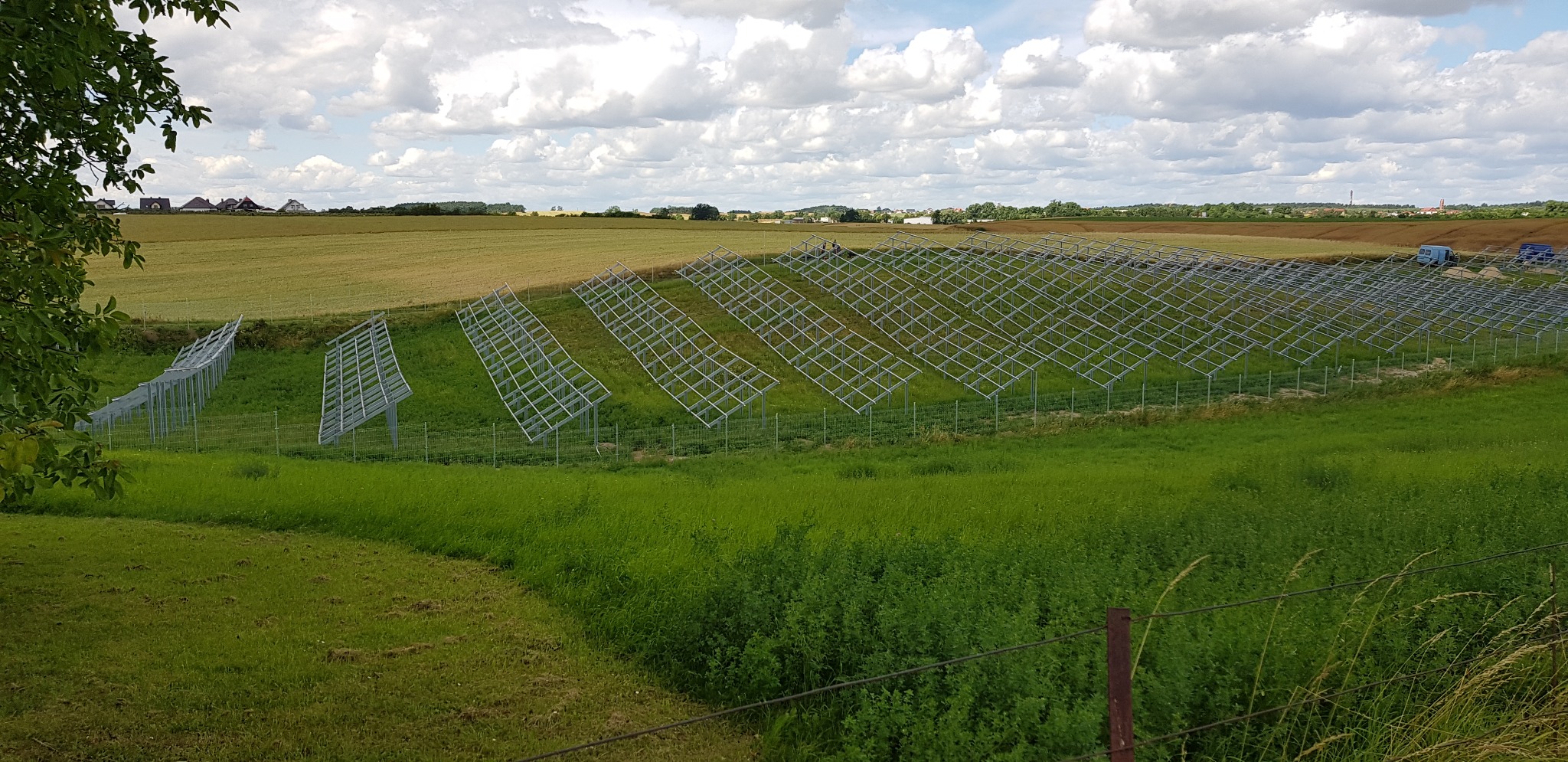 Rozległy widok na konstrukcje wsporcze pod panele słoneczne na zielonym polu, z widocznymi w oddali zabudowaniami i polami uprawnymi pod błękitnym niebem z chmurami.