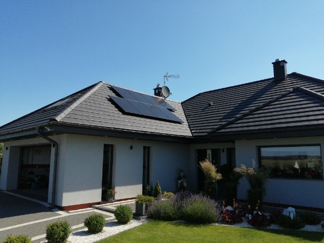 Dom jednorodzinny z szarą dachówką i panelami słonecznymi na dachu, antena satelitarna, zadbany ogród z lawendą i ozdobnymi trawami.