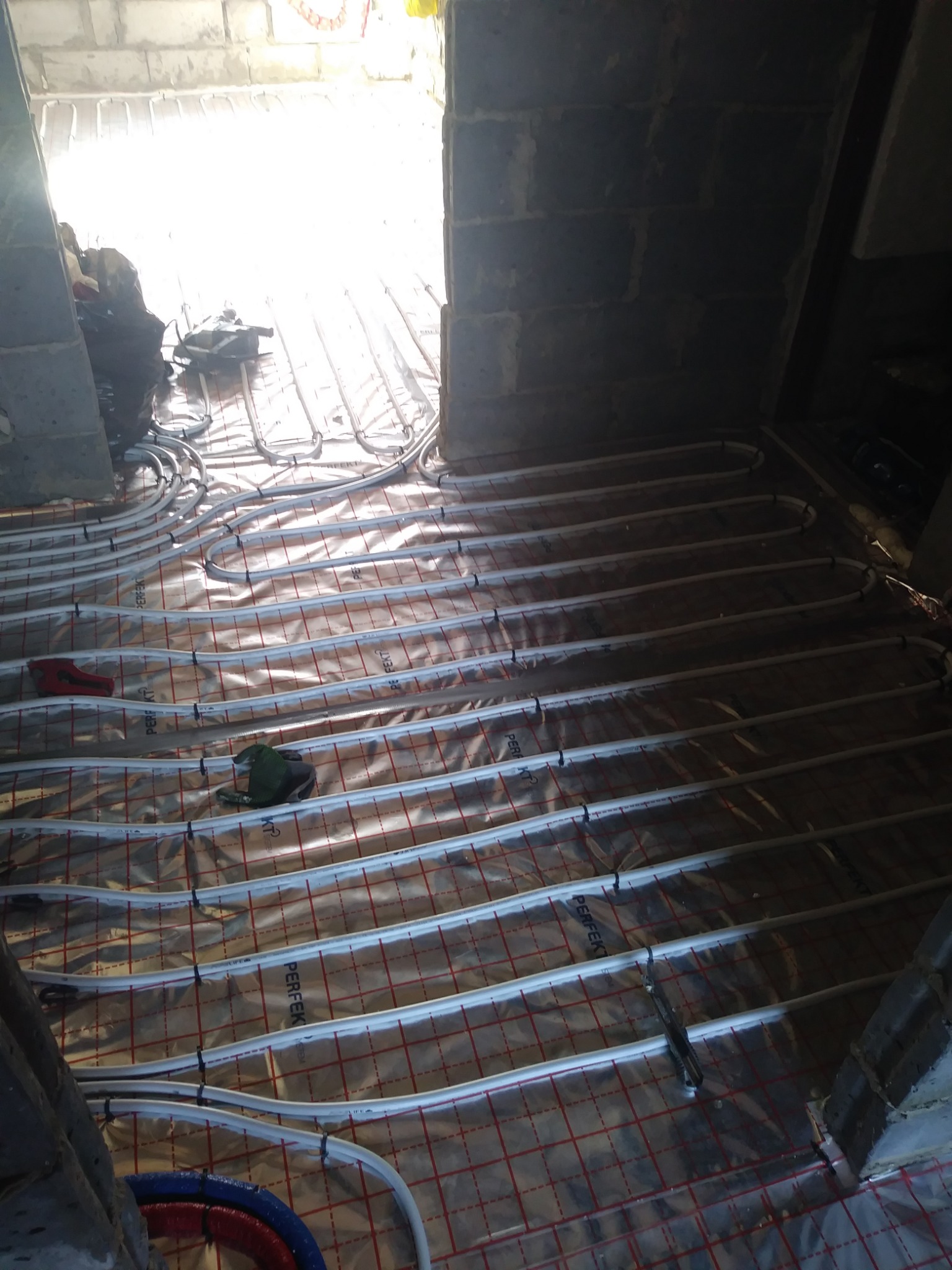 Instalacja ogrzewania podłogowego na folii z czerwoną siatką, widoczne białe rurki przytwierdzone do podłoża, w tle nieotynkowane ściany z pustaków