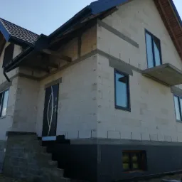 Niedokończony dom murowany z betonu komórkowego, widoczne okna z grafitowymi ramami, czarna stolarka dachowa i ocieplenie fundamentów, schody wejściowe w trakcie budowy.