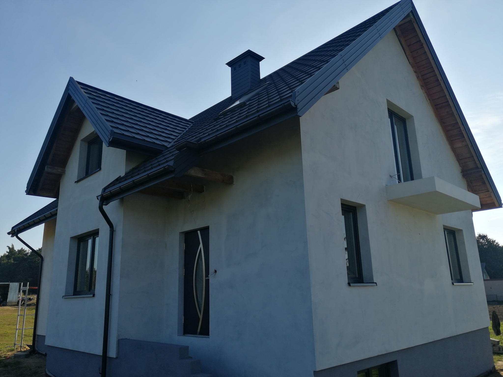 Nowoczesny dom murowany z ciemną dachówką, jasną elewacją i charakterystycznym kominem w słoneczny dzień.