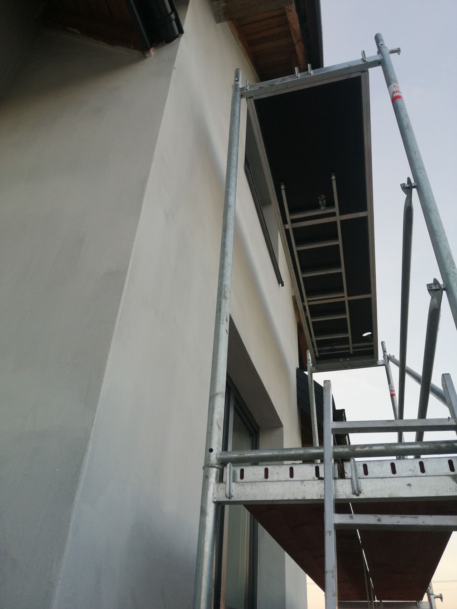 Świeżo otynkowana, biała ściana domu jednorodzinnego widziana z dołu, z fragmentem rusztowania i oknem w tle.