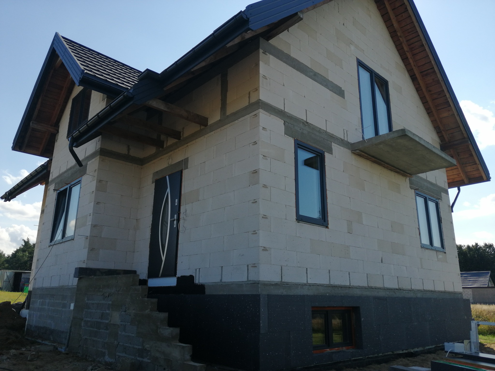 Niedokończony dom murowany z betonu komórkowego, widoczne okna z grafitowymi ramami, czarna stolarka dachowa i ocieplenie fundamentów, schody wejściowe w trakcie budowy.