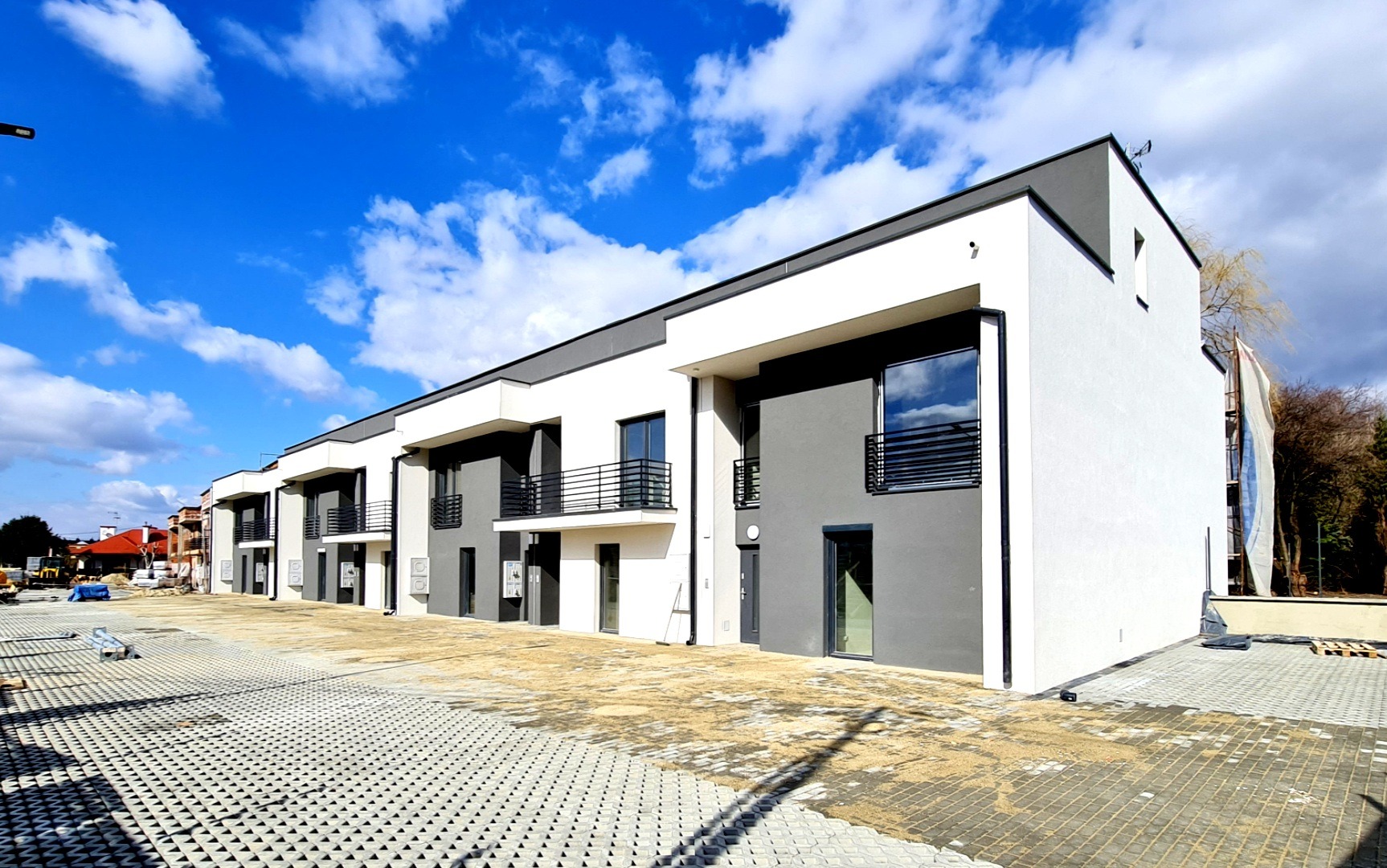 Nowoczesny szeregowiec z szaro-białą elewacją, balkonikami z czarną balustradą i kostką brukową na podjeździe, widok z poziomu gruntu w słoneczny dzień.