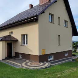 Dom jednorodzinny w Spytkowicach po renowacji elewacji, widoczny z zewnątrz, z brązowym dachem i beżową elewacją, z dekoracyjnym wzorem z kostki brukowej przed wejściem.