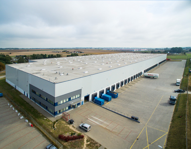 Rozległy, nowoczesny magazyn z rampami załadunkowymi, widok z lotu ptaka, w tle pola uprawne i pochmurne niebo.