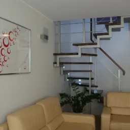 Nowoczesne wnętrze domu z drewnianymi schodami na metalowej konstrukcji, jasną sofą i abstrakcyjnym obrazem z czerwonymi okręgami na ścianie.