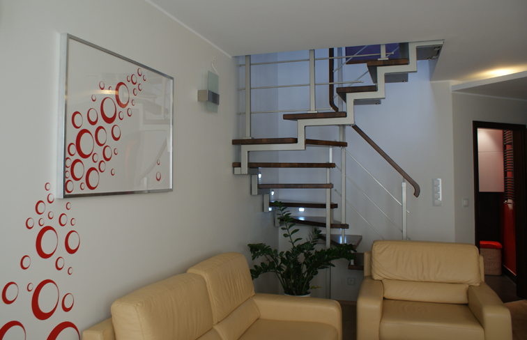 Nowoczesne wnętrze domu z drewnianymi schodami na metalowej konstrukcji, jasną sofą i abstrakcyjnym obrazem z czerwonymi okręgami na ścianie.