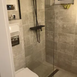 Nowoczesna łazienka z kabiną prysznicową typu walk-in z czarną armaturą, wiszącą toaletą i szarymi płytkami imitującymi kamień.