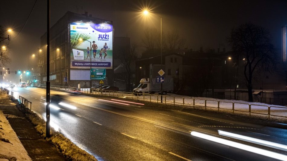 Nocny widok na oświetloną ulicę z billboardem reklamowym na budynku w tle, z efektem rozmycia świateł przejeżdżających samochodów.