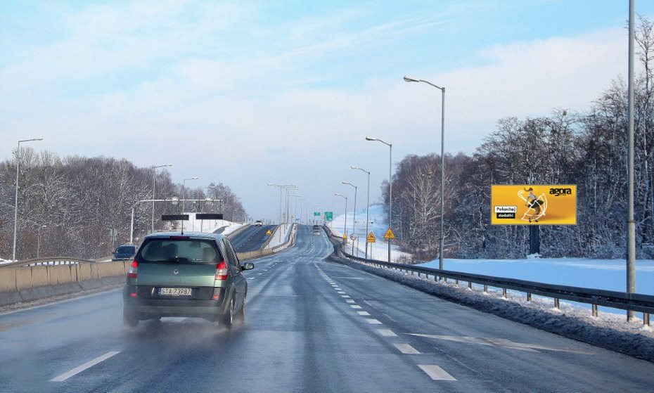 Billboard 'Agora' z żółtym tłem i postacią w ruchu, widoczny z jadącego samochodu na autostradzie w zimowej scenerii.