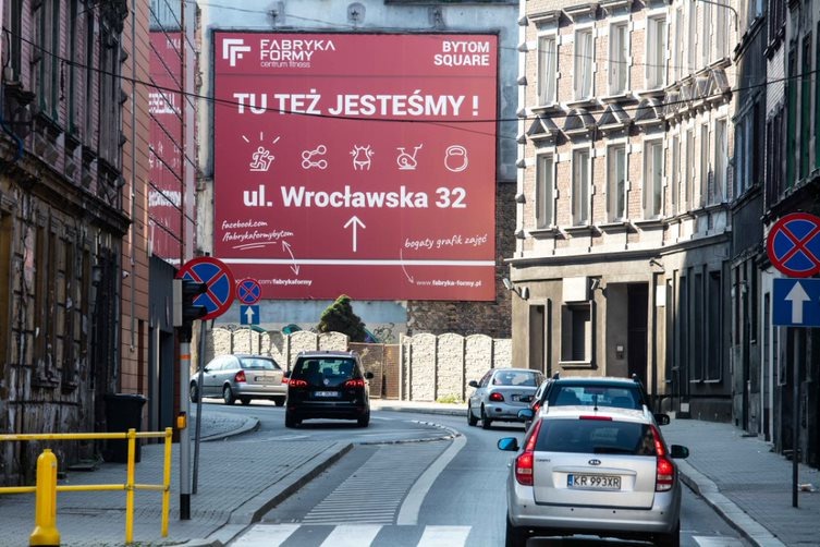 Billboard reklamowy centrum fitness 'Fabryka Formy' umieszczony na ścianie budynku przy ulicy Wrocławskiej 32, widoczny z perspektywy ulicy z przejeżdżającymi samochodami.
