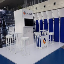 Brand Creation Sp.j - Biało-niebieskie stoisko targowe LOT AMS z logo, ekranem i grafiką samolotu w tle, wyposażone w białe stoliki i hokery.