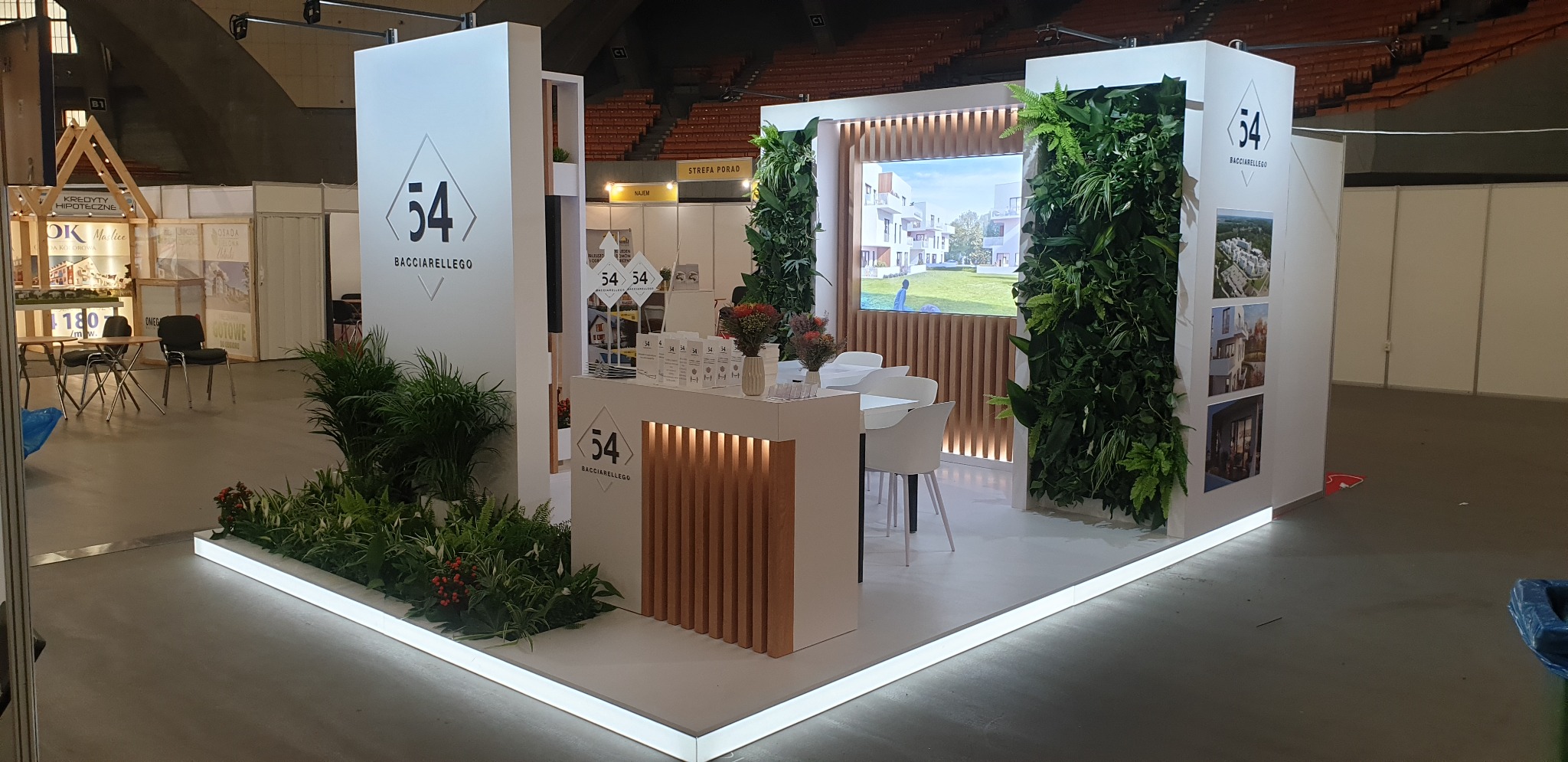 Stoisko targowe z białymi meblami, logo firmy Bacciarellego, pionowymi ogrodami i drewnianymi akcentami, oświetlone od dołu.