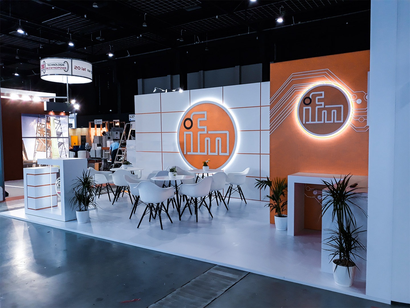 Aranżacja stoiska targowego z okrągłymi, podświetlanymi logotypami, białymi krzesłami i stołami kawowymi oraz roślinami doniczkowymi, utrzymana w pomarańczowo-białej kolorystyce.