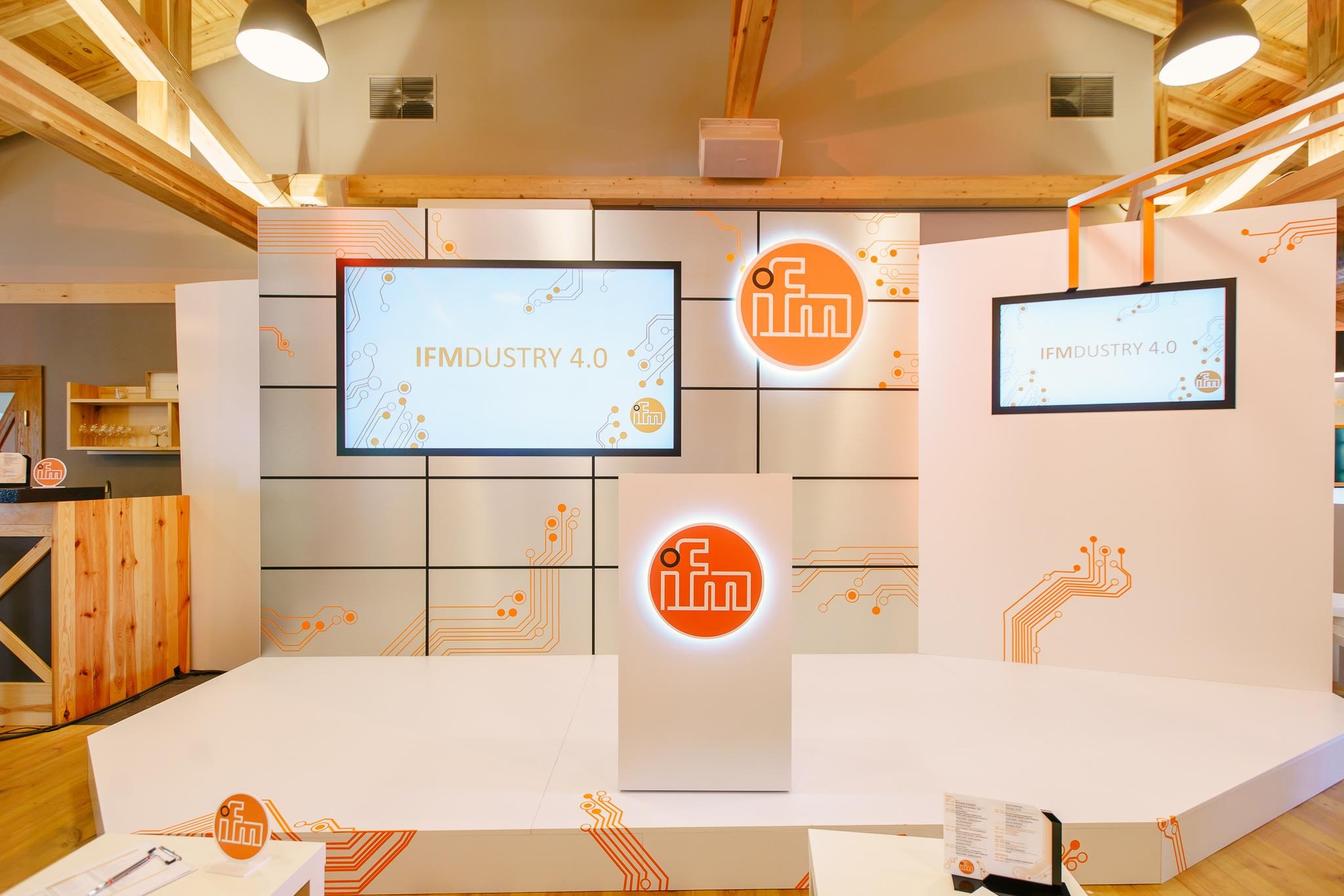 Scena z dwoma ekranami wyświetlającymi 'IFMDUSTRY 4.0' i logo, na białym tle z pomarańczowymi detalami przypominającymi obwody elektroniczne, oświetlona ciepłym światłem.