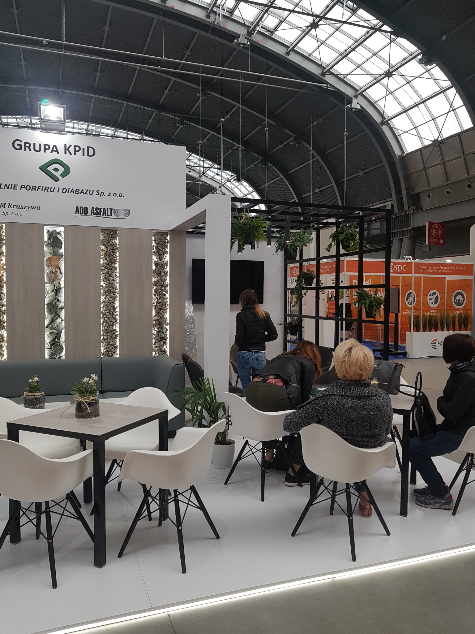 Stoisko targowe firmy Grupa KPID z panelami dekoracyjnymi z kamienia naturalnego, stoliki kawowe z białymi krzesłami i roślinami, widoczni zwiedzający.